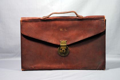 Mallette à rabat rectangulaire en cuir brun. Elle possède un fermoir en métal dorée et une poignée sur le dessus. On retrouve les lettres W.L.( Wilfrid Laurier) en or inscrit sur le devant.