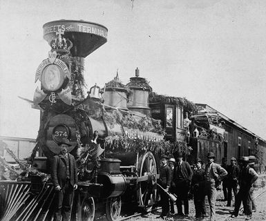 Photo d'une locomotive à cheminer, décorer avec des guirlandes et les armoiries de la reine Victoria pour son Jubilés. La locomotive est attachée à plusieurs wagons. Un groupe d'hommes se trouve devant l'engin.