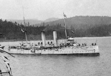 Photo en noir et blanc d'un navire en métal à deux mats à chaque extrémité et  à deux cheminées au centre, traînant derrière lui un petit bateau de bois. Derrière le bateau on observe une grande forêt qui caractérise le port de Prince Rupert en 1914.